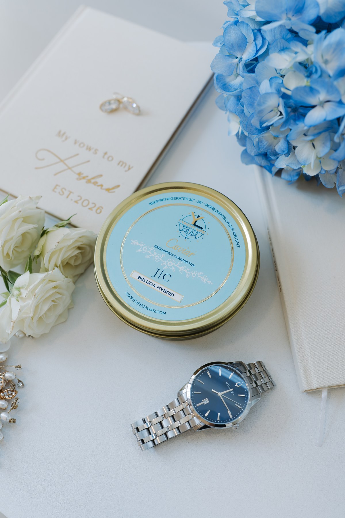 A Yacht Life Caviar tin with a custom pale blue label reading 'Exclusively curated for J | C, Beluga Hybrid', photographed on a wedding morning beside the rings, a vow book, blue hydrangeas and white roses